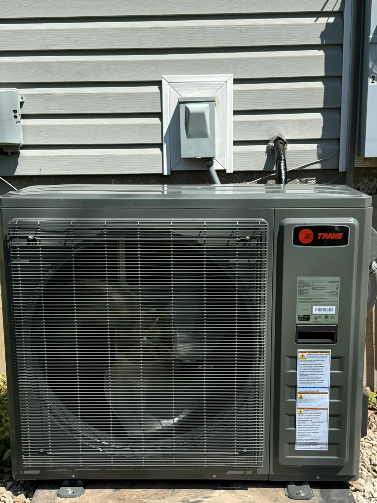A gray outdoor unit on a concrete pad against light horizontal siding, with a disconnect box mounted above it.