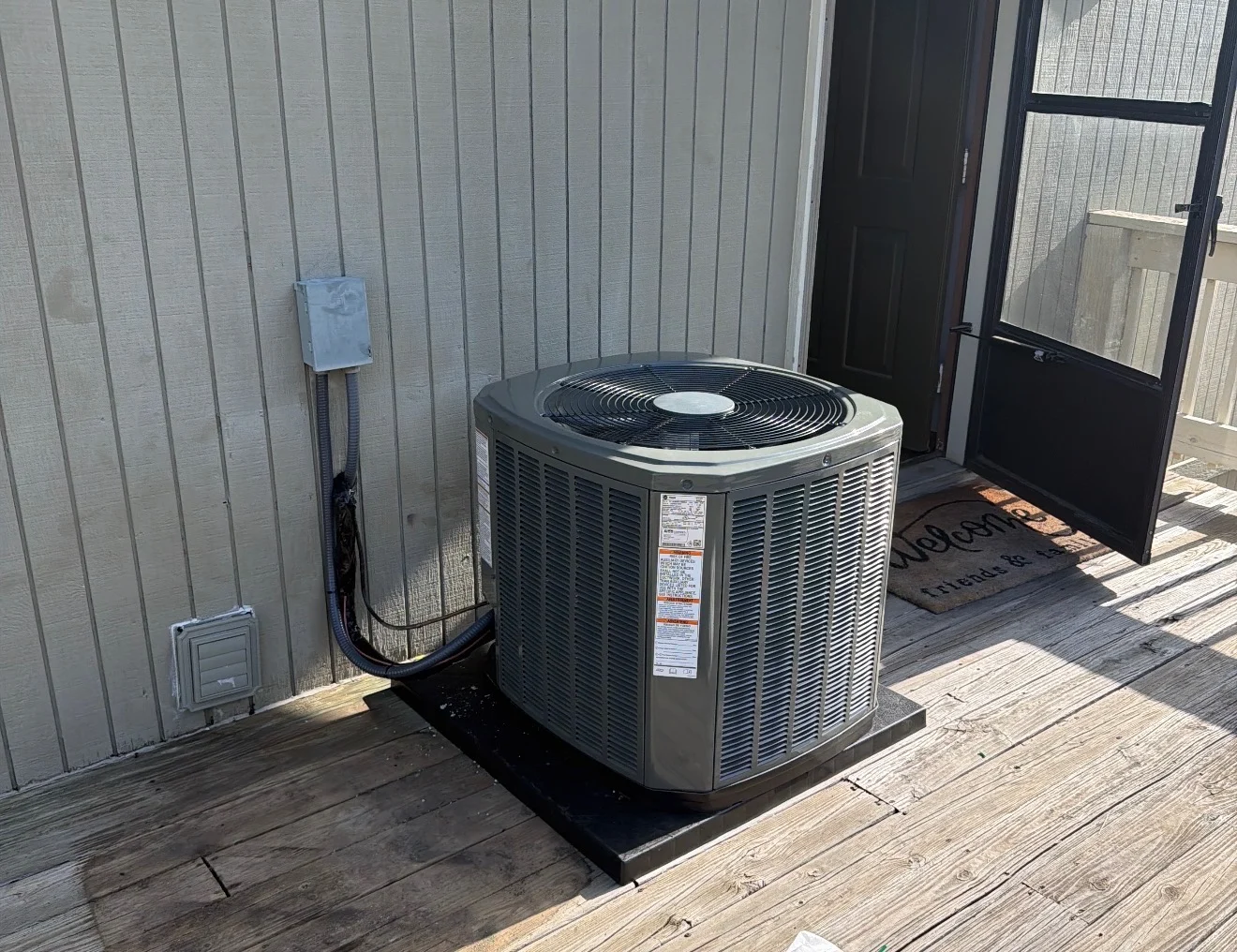 A clean outdoor condensing unit installed on a pad on a wood deck, with the disconnect and line set finished against the wall.